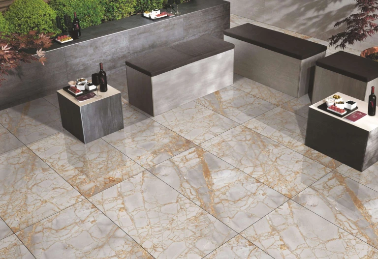 A modern outdoor patio features marble-patterned tiles, gray and brown benches, and small tables with wine bottles and glasses. Surrounded by greenery, the stylish space is as inviting as a soothing stream, blending nature with design under the careful hands of expert construction.