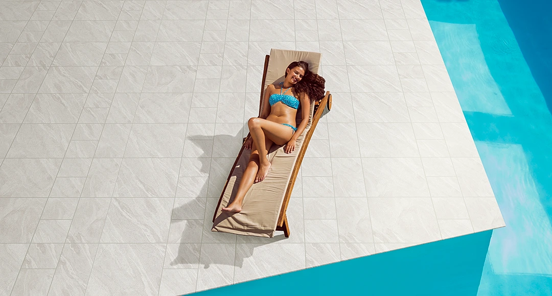 A woman in a blue swimsuit relaxes on a lounge chair by the pool, basking in the sunlight streaming onto the white tiled surface.